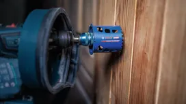 Une scie cloche fixée à une perceuse électrique effectue une découpe circulaire dans un mur en bois.